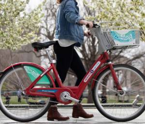 A woman with a jean jacket and black jeans walking a B-cycle bike with a circuit media green graphic on the basket